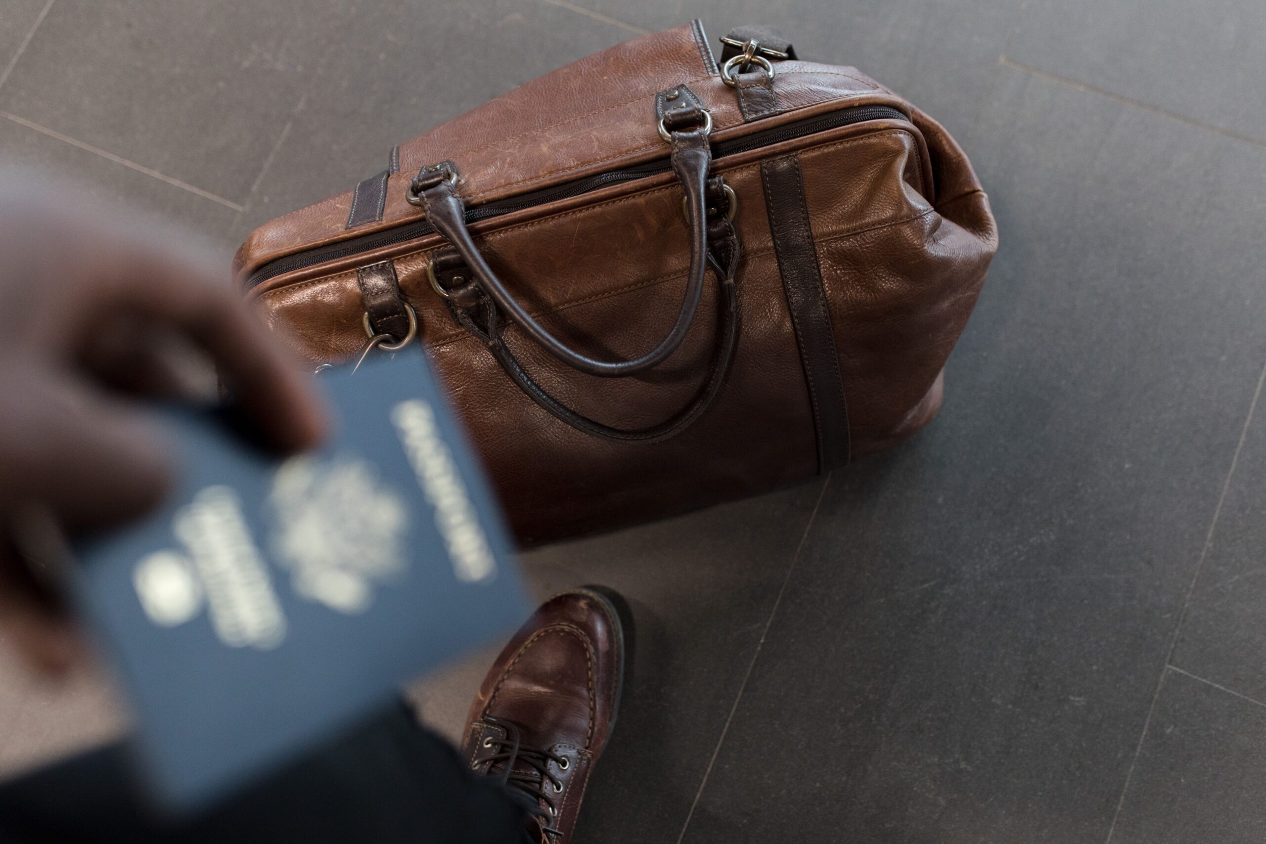 Brown leather holdall bag on the floor with a passport held nearby, travel setting