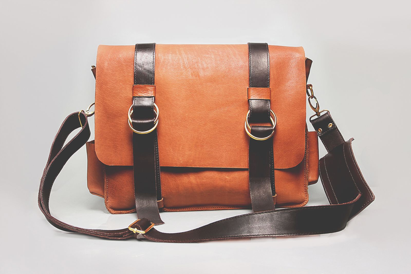 A brown leather satchel-style briefcase with contrasting dark leather straps and metal buckles, studio shot