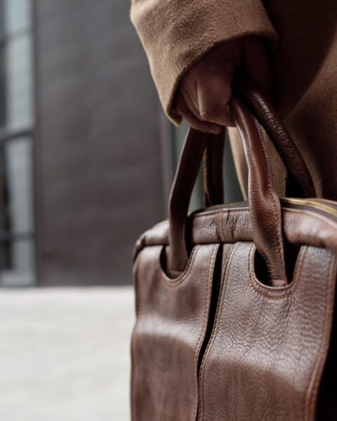 Close-up of a hand carrying a premium brown leather briefcase against a modern city backdrop