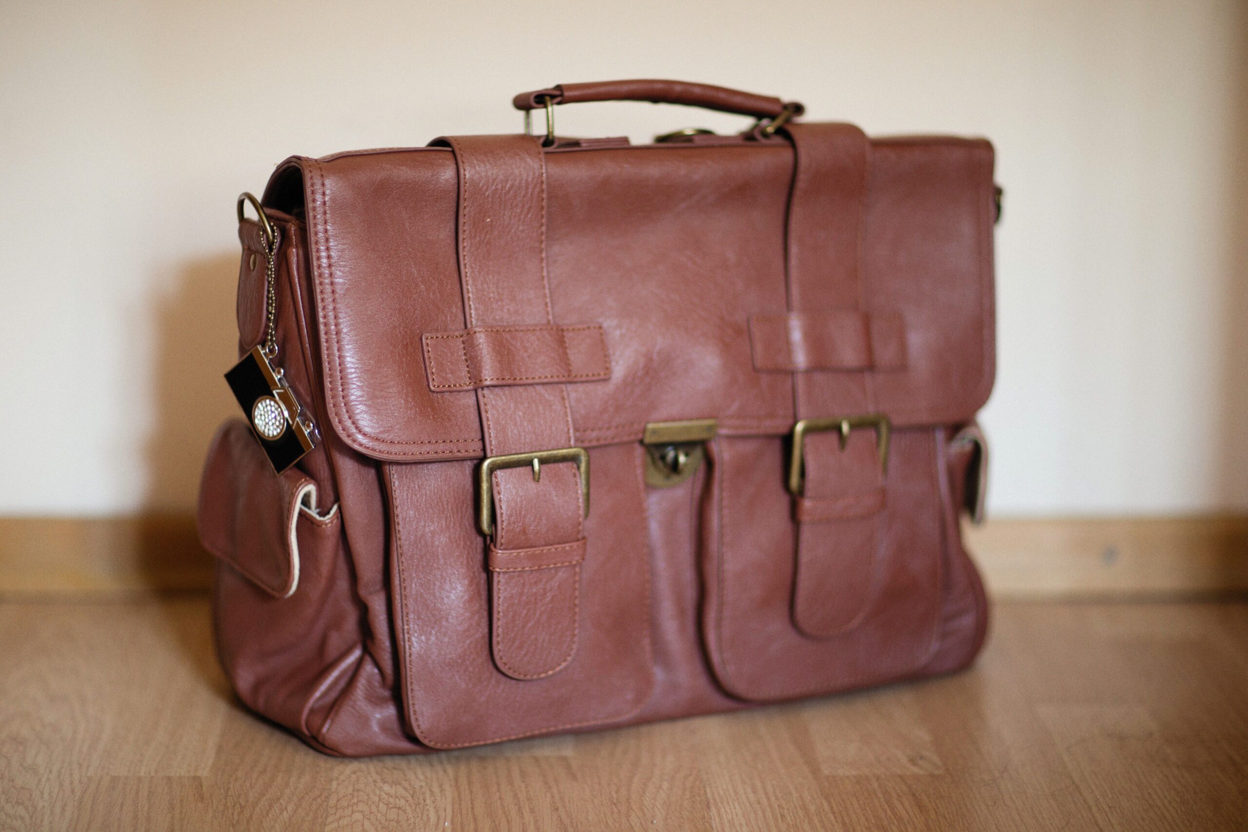 Tan leather satchel with front buckles resting on a wooden floor against a warm plaster wall