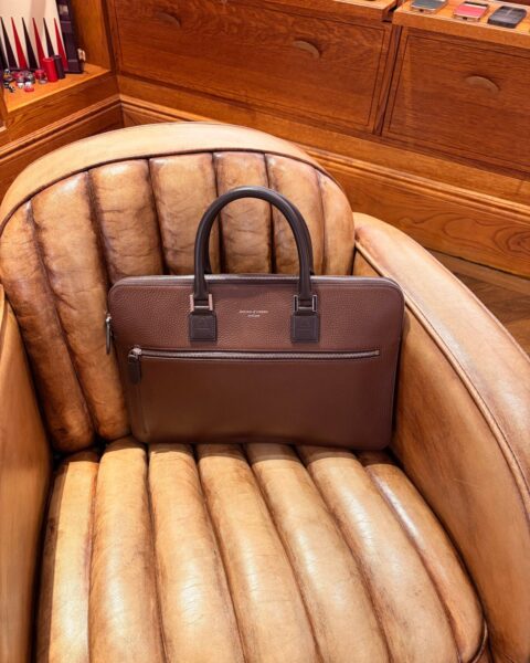 An Aspinal of London briefcase in brown pebble-grain leather with silver hardware and a full-width front zip pocket, on a worn leather club chair in the Regent Street shop