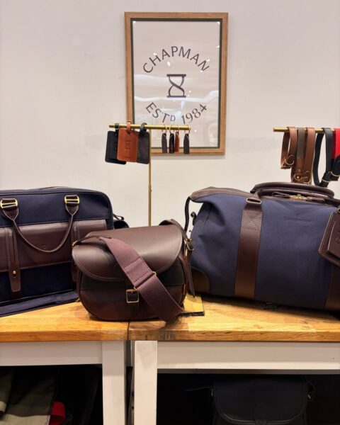 Chapman's King's Road pop-up: a navy canvas briefcase, a brown leather cartridge bag and the navy canvas Weekender on a wooden table, under a framed "Chapman Est'd 1984" sign