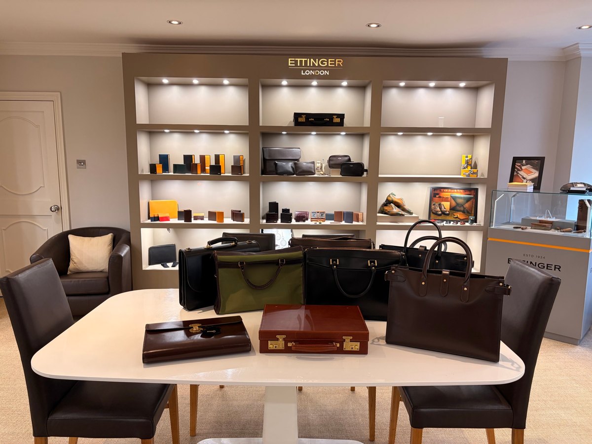 Ettinger's Putney showroom: briefcases, totes, a flap-over case and a cognac attaché case on a white table, in front of lit shelves of wallets and small leather goods