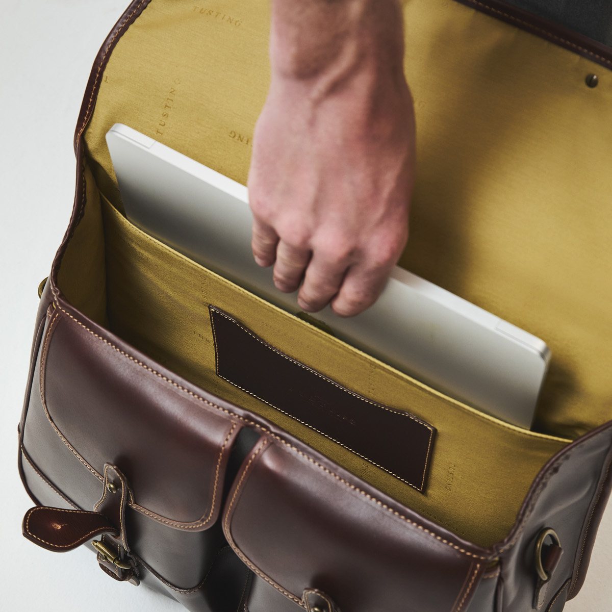 Open Tusting Leather Clipper in Sundance Floodlight leather from above, showing the mustard jacquard lining with the Tusting name woven in, a leather patch pocket and a laptop being lifted out